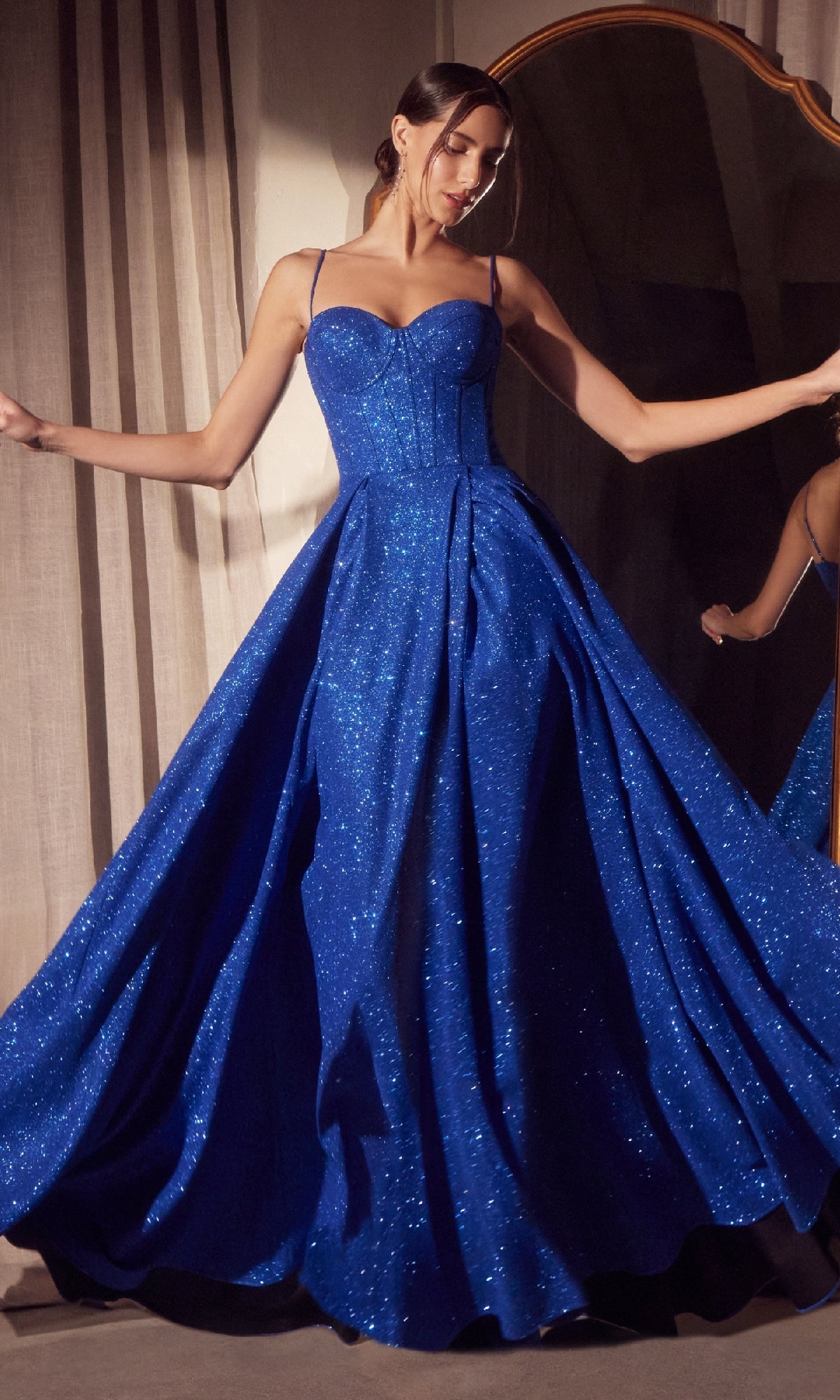 A woman poses in front of a large mirror wearing the Amoris T706 Lace-Up Long Glitter Prom Ball Gown by Cinderella Divine, a sparkling royal blue dress featuring thin straps and a fitted bodice.